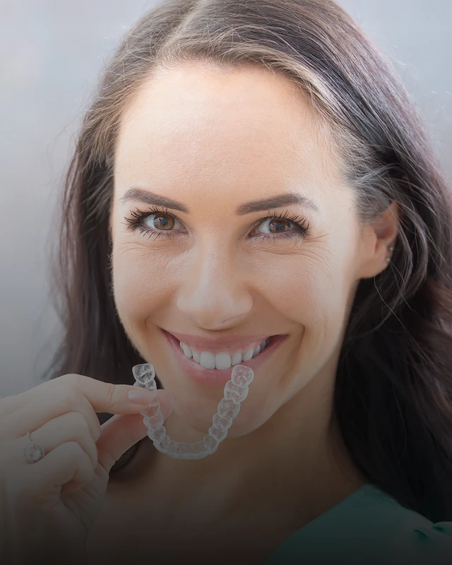 A woman smiling wide and holding up her Invisalign clear aligners